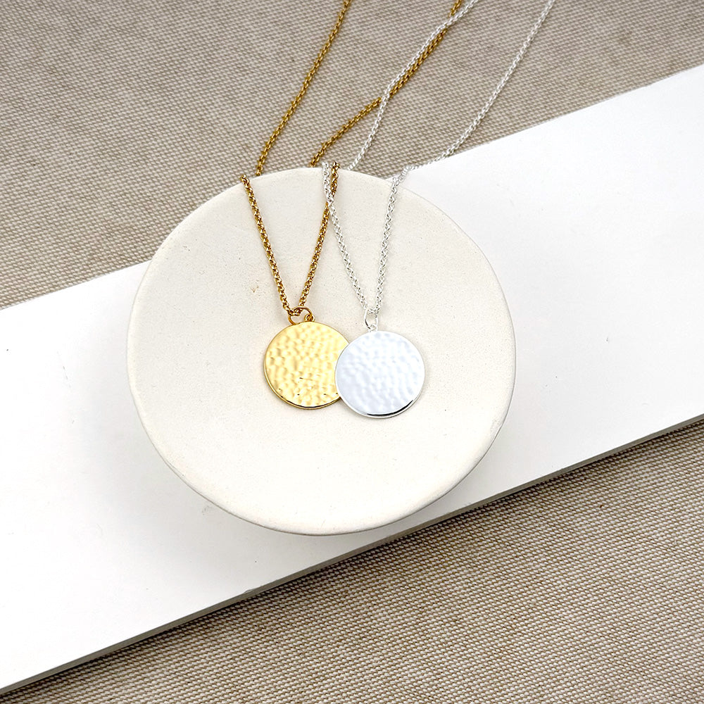 Two minimalist round pendant necklaces displayed on a white ceramic dish. One necklace has a textured gold pendant on a gold chain, and the other has a textured silver pendant on a silver chain, set against a neutral beige fabric background.