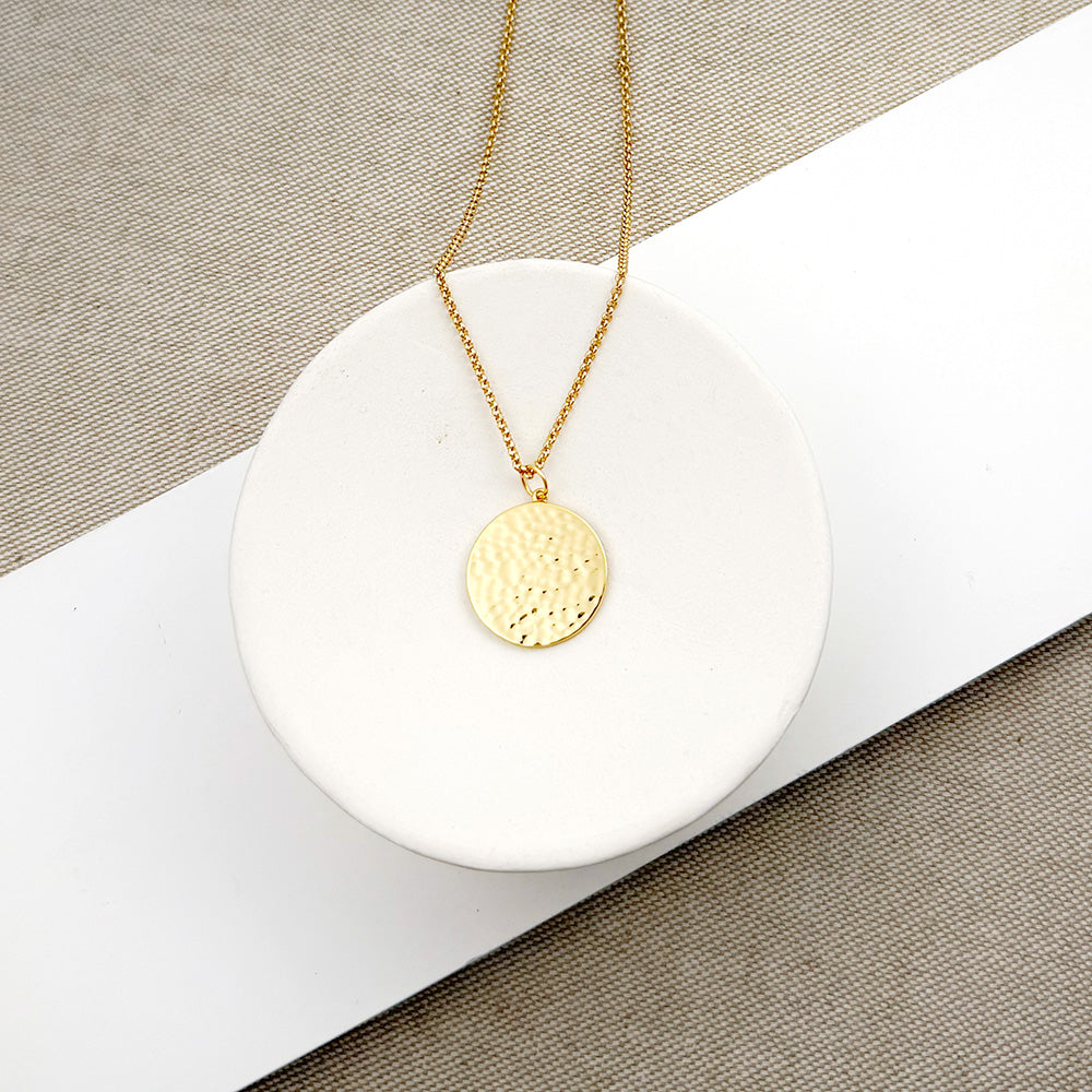 A gold chain necklace with a textured, hammered circular pendant, displayed on a white ceramic dish over a neutral beige and white background.