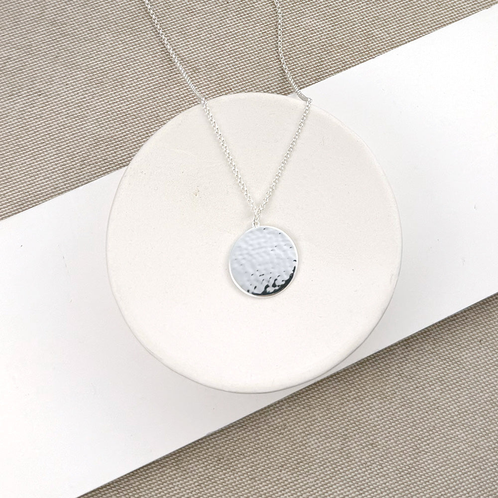 A silver necklace with a textured, hammered circular disc pendant, displayed on a white circular stand over a beige woven fabric background.