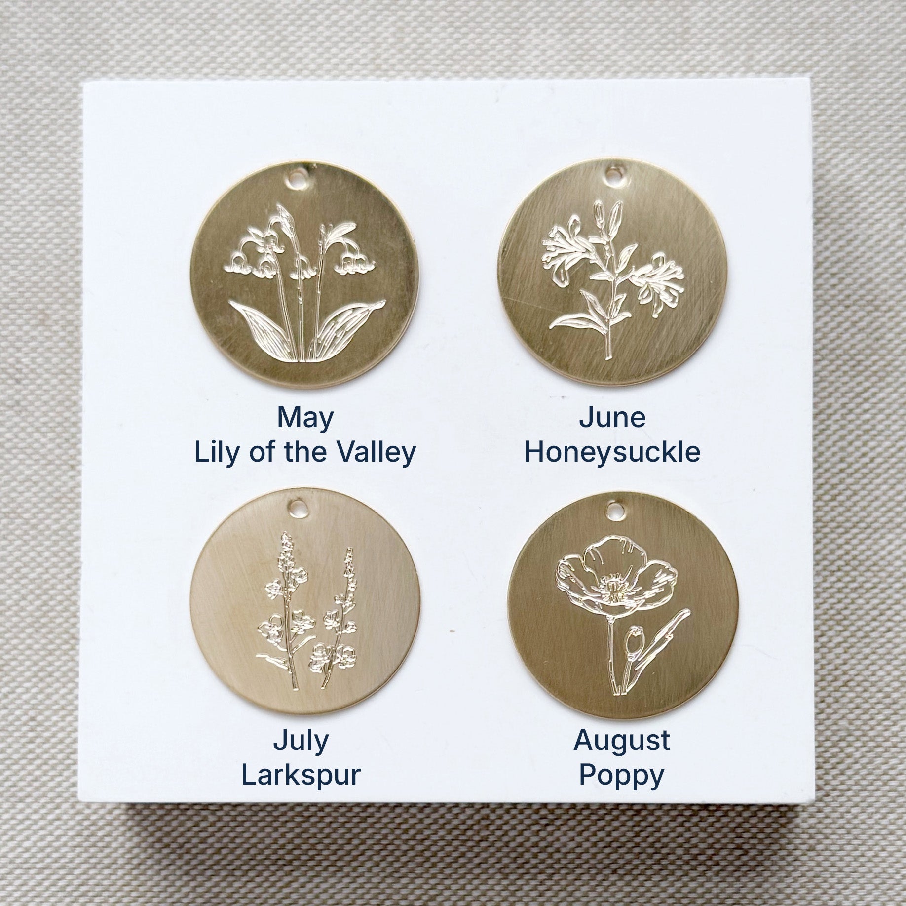 Four gold floral charms on a white card with month and flower name labels, on a textured beige background.