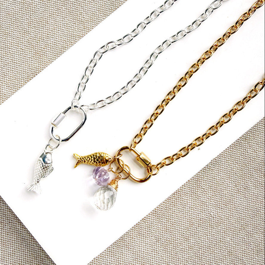 Two delicate chain necklaces displayed on a white rectangular card against a beige textured fabric background. One necklace is silver with a fish charm and a small clear crystal pendant.