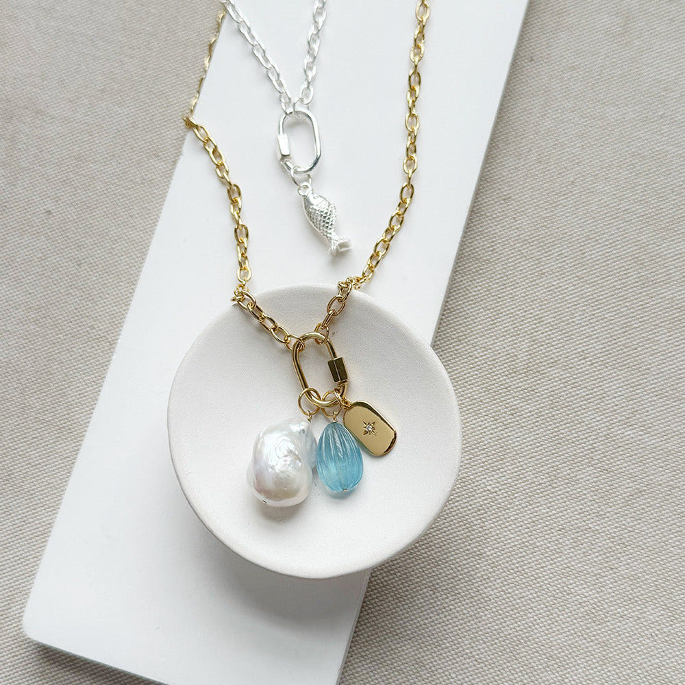 A gold chain necklace with a fish-shaped charm and three dangling charms (a white pearl, a faceted light blue stone, and a small gold tag) displayed on a white dish over a neutral beige fabric background.