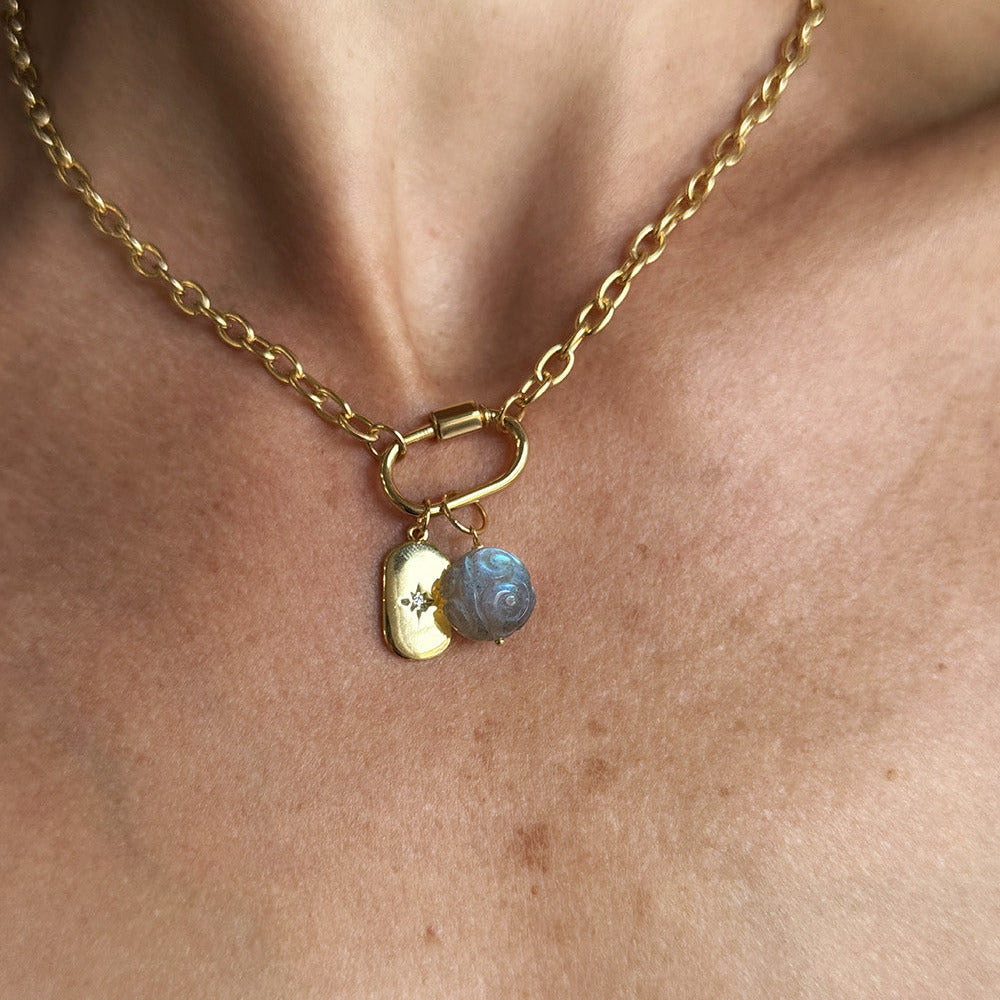 A close-up of a gold chain necklace worn on a person's neck, featuring a small gold tag charm with a star and a round carved labradorite charm.