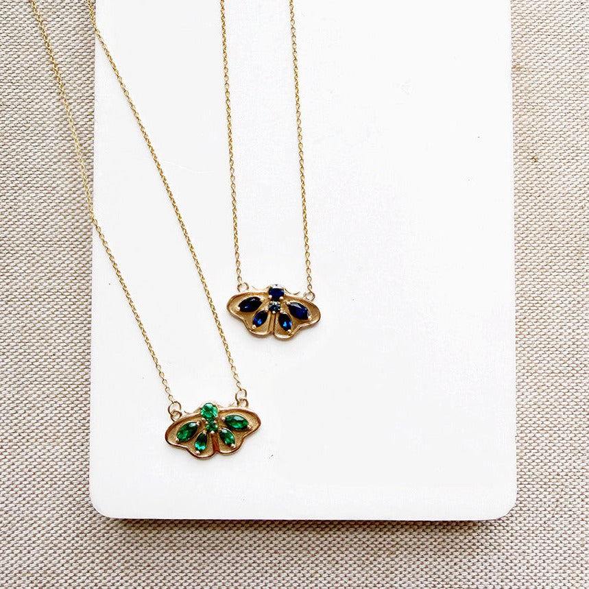 Two delicate gold chain necklaces with butterfly-shaped pendants, one featuring green stones and the other blue stones, displayed on a white card over a beige textured fabric background.