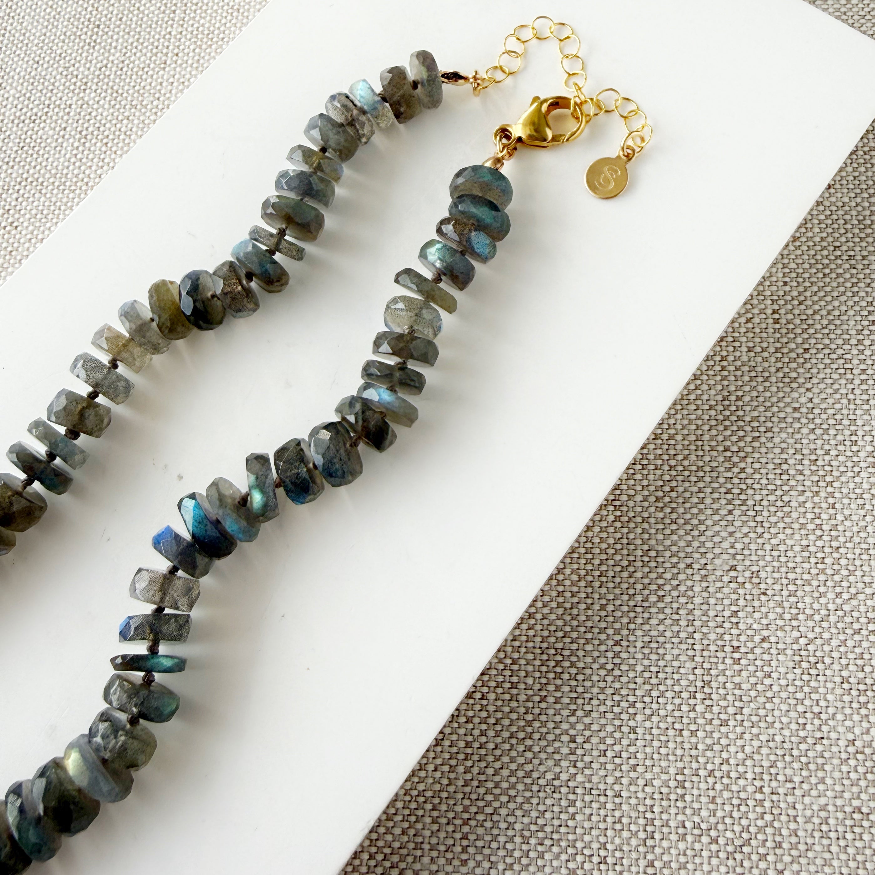 Clasp detail of a necklace with labradorite gemstones on a white dish against a beige fabric background