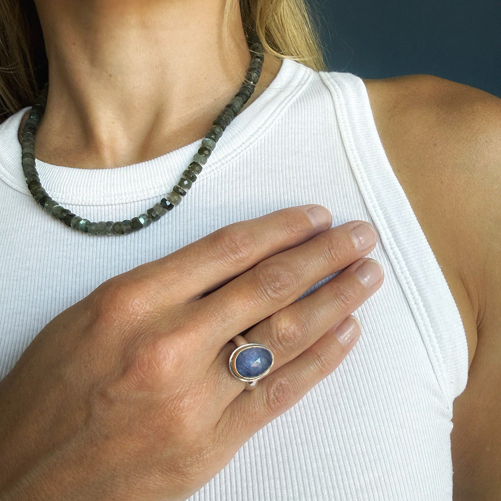 A close-up of a person wearing a silver ring with a tanzanite cabochon stone, paired with a dark green beaded necklace and a white ribbed tank top.