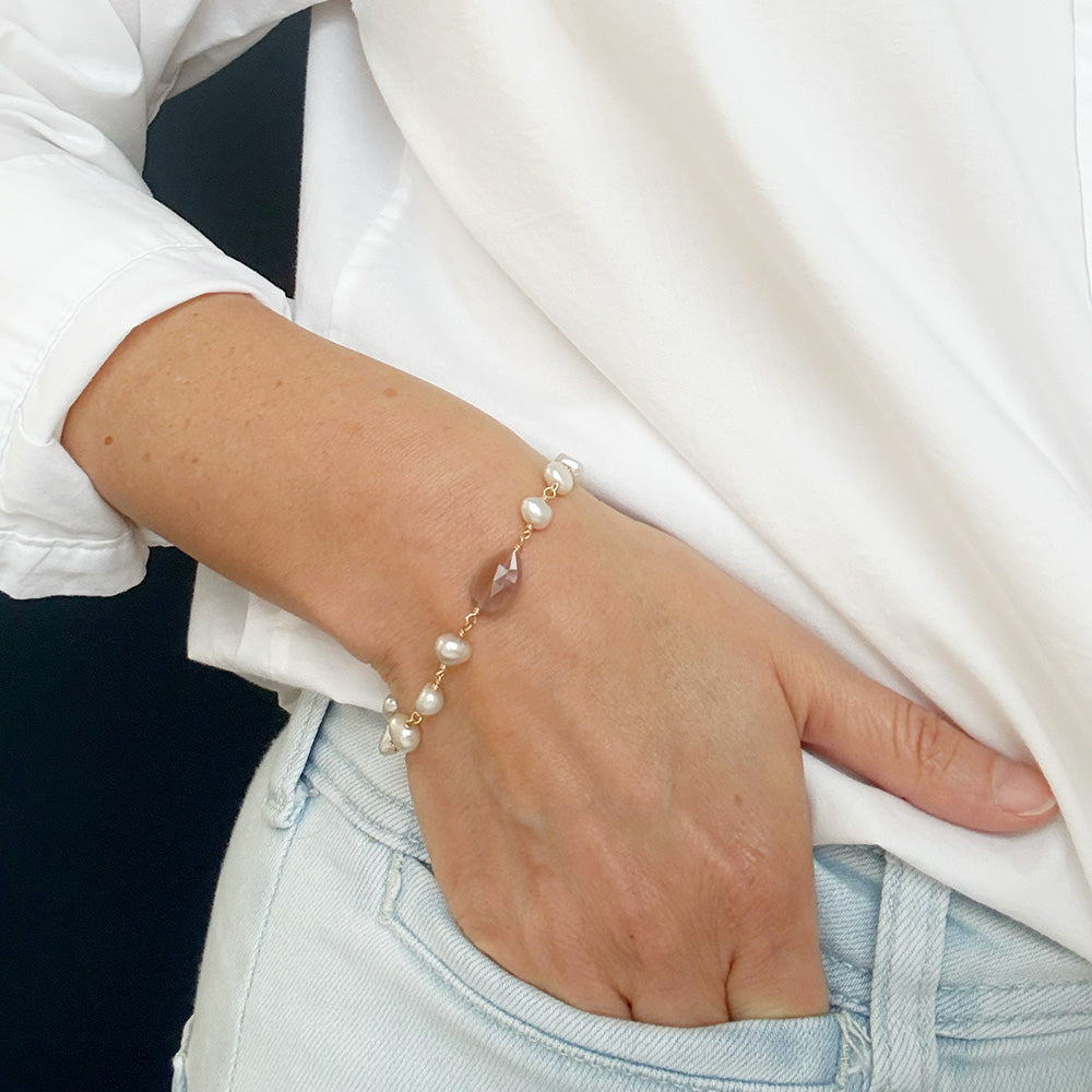 A delicate bracelet with white pearls and a central coffee moonstone teardrop stone, worn on a person's wrist.