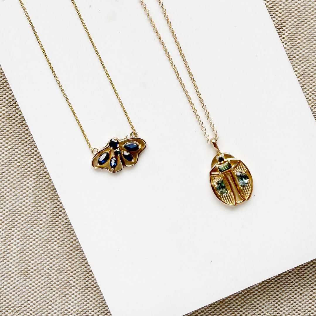 Two gold necklaces with butterfly and beetle pendants on a white surface