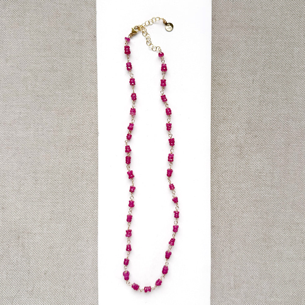 A necklace featuring pink sapphires linked by gold-toned metal, displayed on a white rectangular card over a textured beige fabric background.