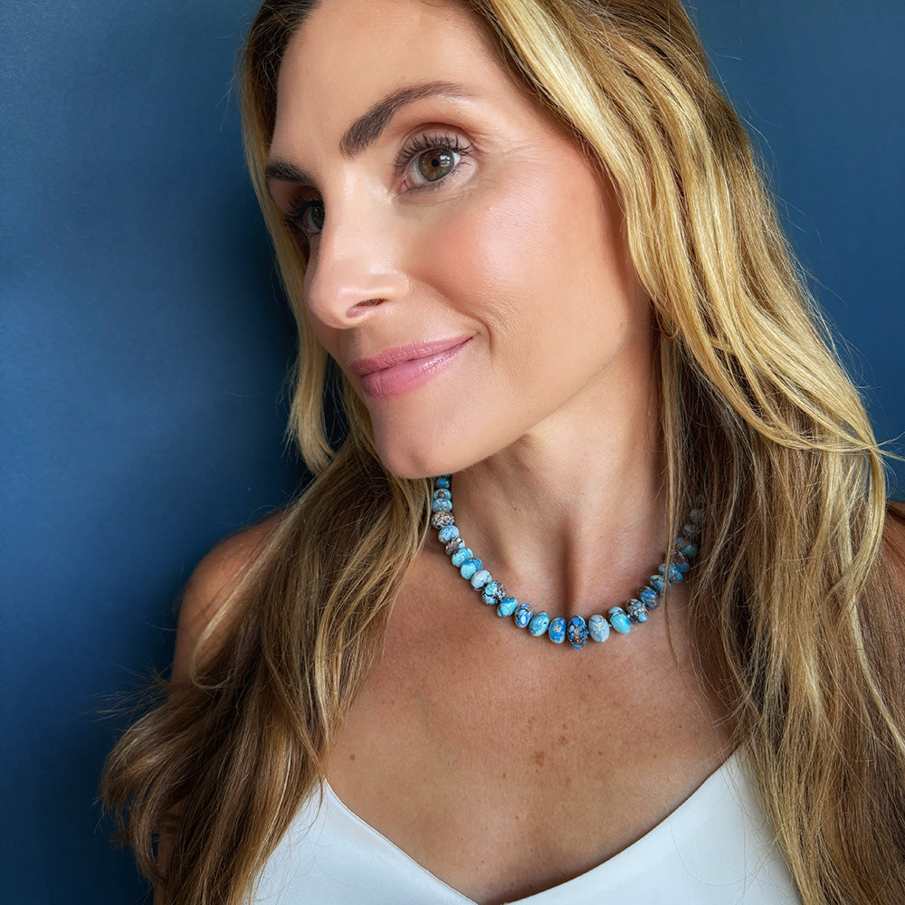 A woman with blonde hair wearing a turquoise beaded necklace, photographed against a dark blue background.