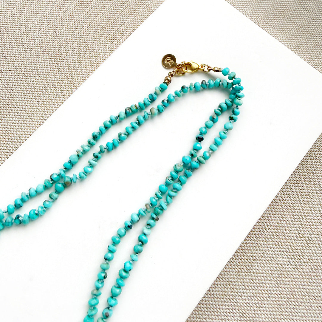 Turquoise beaded necklace with a gold clasp on a white card with a beige fabric background