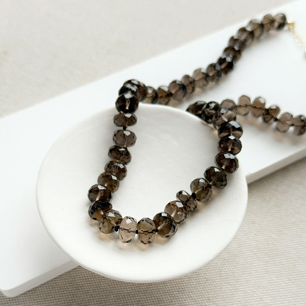 A knotted necklace of faceted smokey quartz gemstones resting on a white dish. The necklace features a gold-tone lobster clasp and extender chain.