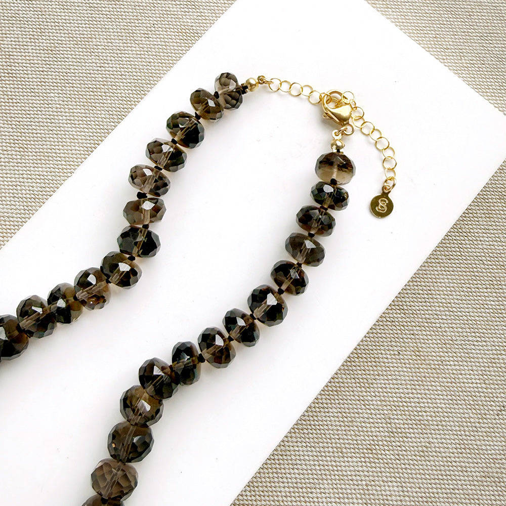 A knotted necklace of faceted smokey quartz gemstones resting on a white dish. The necklace features a gold-tone lobster clasp and extender chain.