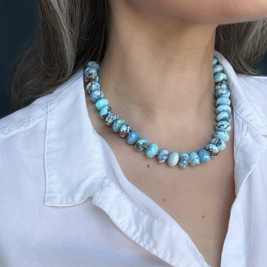 Close up of golden hills necklace with blue beads worn by a person in a white shirt