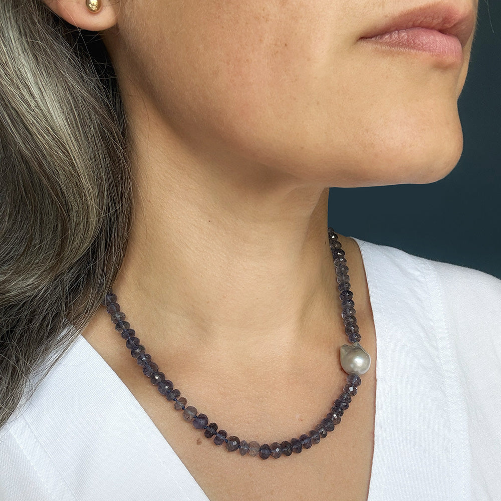A close-up of a woman wearing a beaded necklace made of dark gray faceted beads with a single irregular gray pearl accent, paired with a white top.
