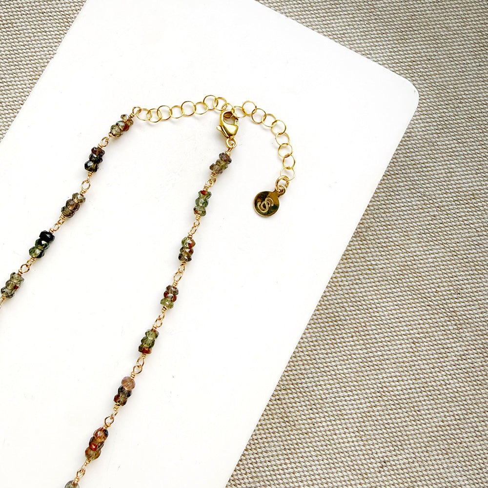 A necklace featuring small green faceted sapphires accented with a lager London Blue Topaz gemstone, displayed on a white rectangular card over a textured beige fabric background.