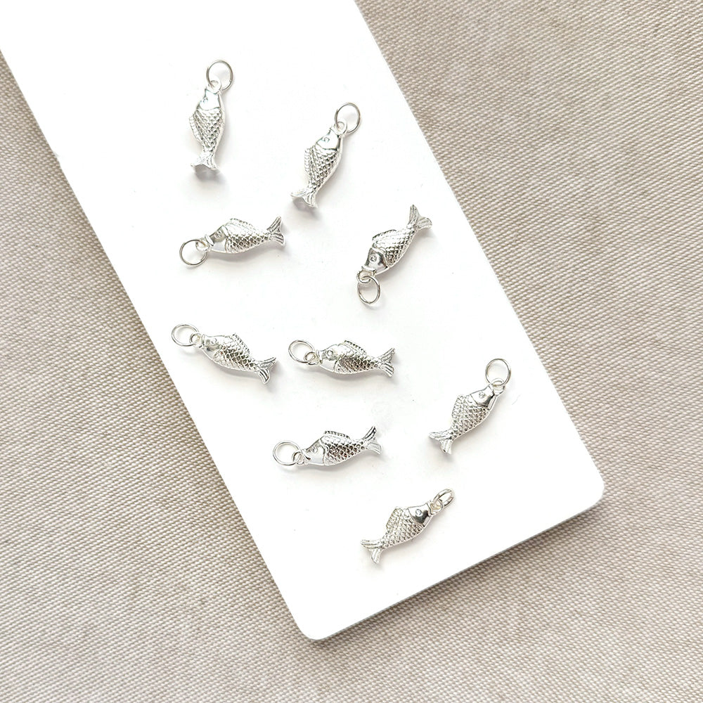 Nine small silver fish-shaped jewelry charms with textured scales and hanging loops, arranged on a white rectangular card over a beige fabric background.