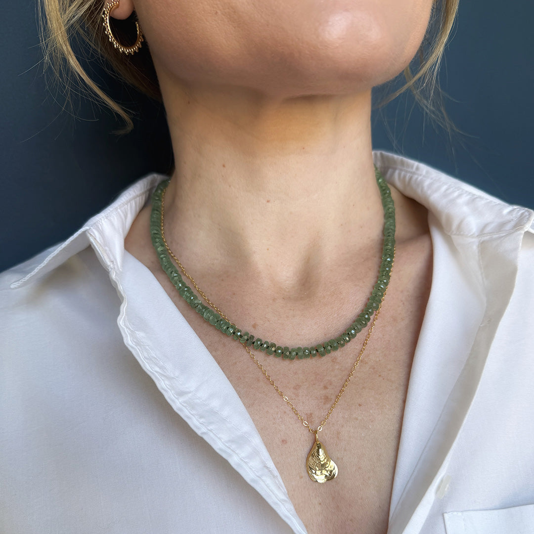 Close-up of a person wearing two necklaces, one green kyanite and one gold with a seashell pendant and a white shirt