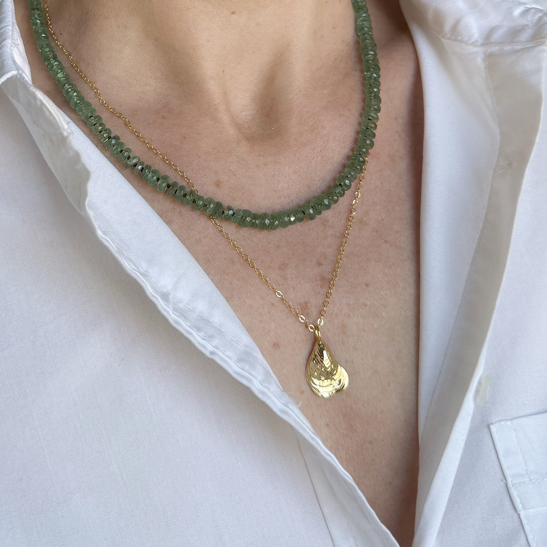 Close-up of a person wearing two necklaces, one green kyanite and one gold with a seashell pendant and a white shirt