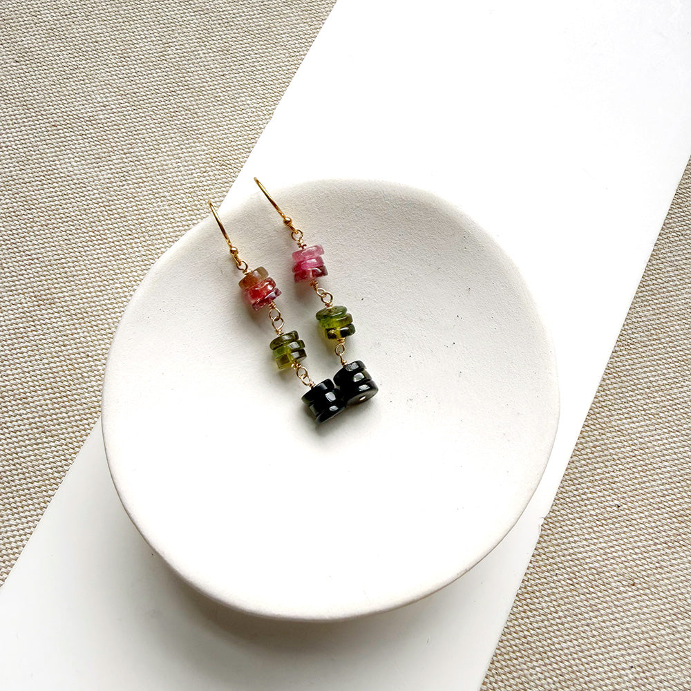 A pair of gold earrings with watermelon tourmaline beads, displayed on a small white ceramic dish over a beige woven fabric background.