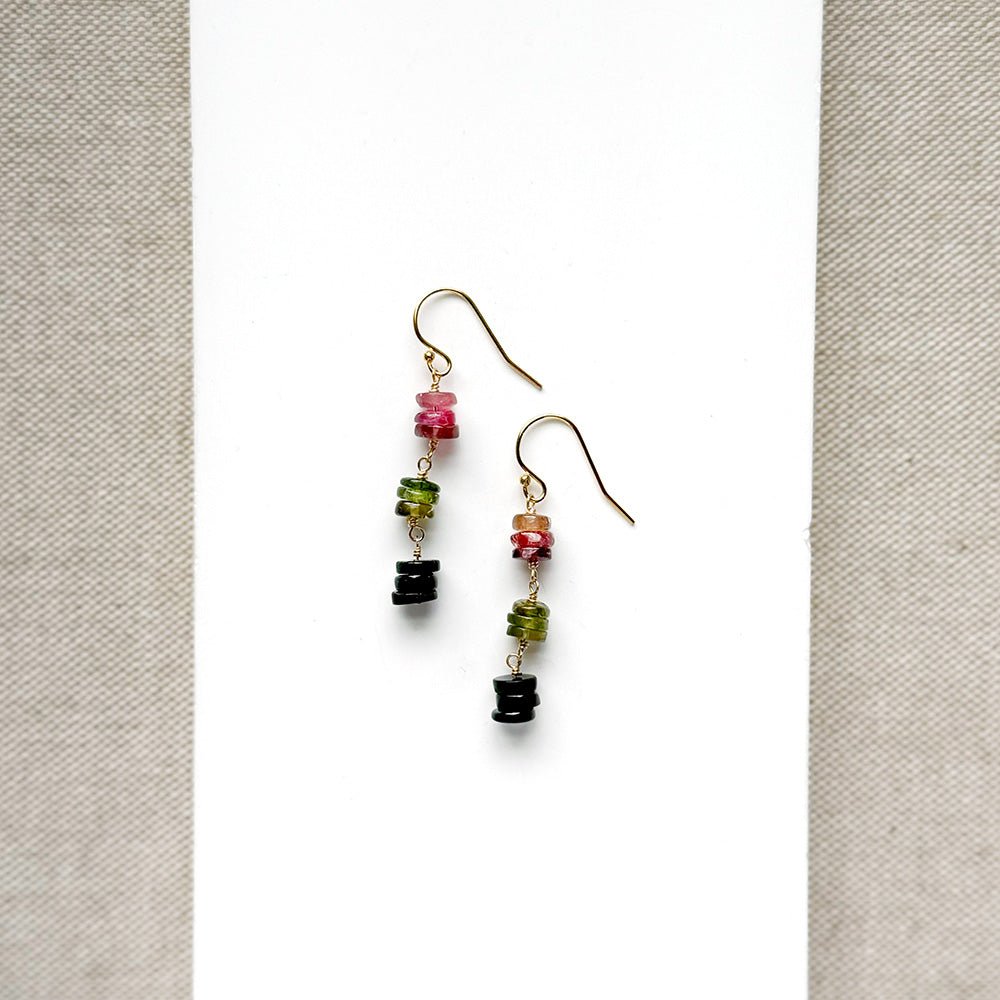 A pair of gold earrings with watermelon tourmaline gemstones, displayed on a white card between textured beige fabric borders.