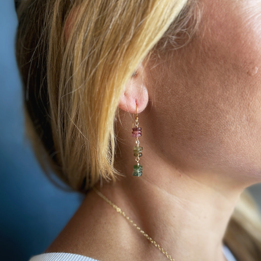 A close-up lifestyle shot of a woman's ear wearing a delicate gold dangle earring watermelon tourmaline gemstone beads, alongside a thin gold chain necklace.