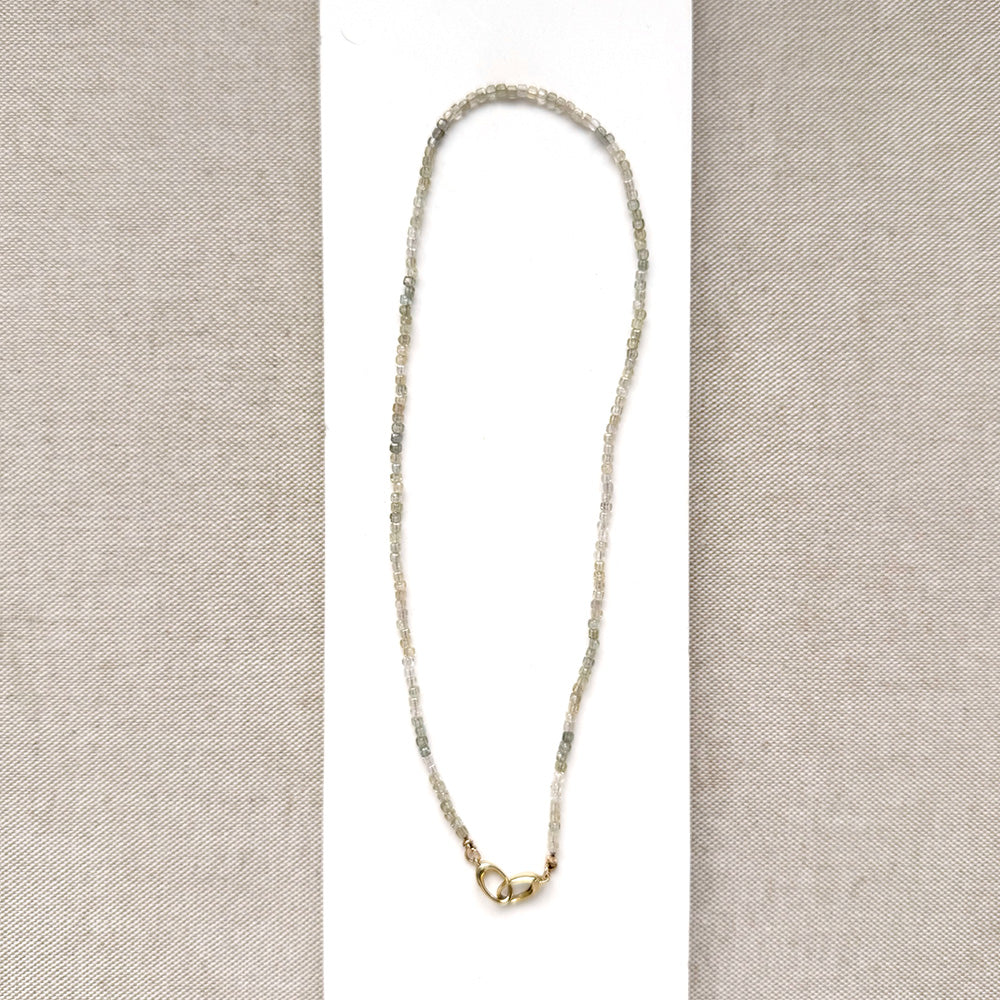 A delicate white zircon gemstone necklace with a gold infinity-style pendant, displayed on a white rectangular card over a beige textured fabric background.
