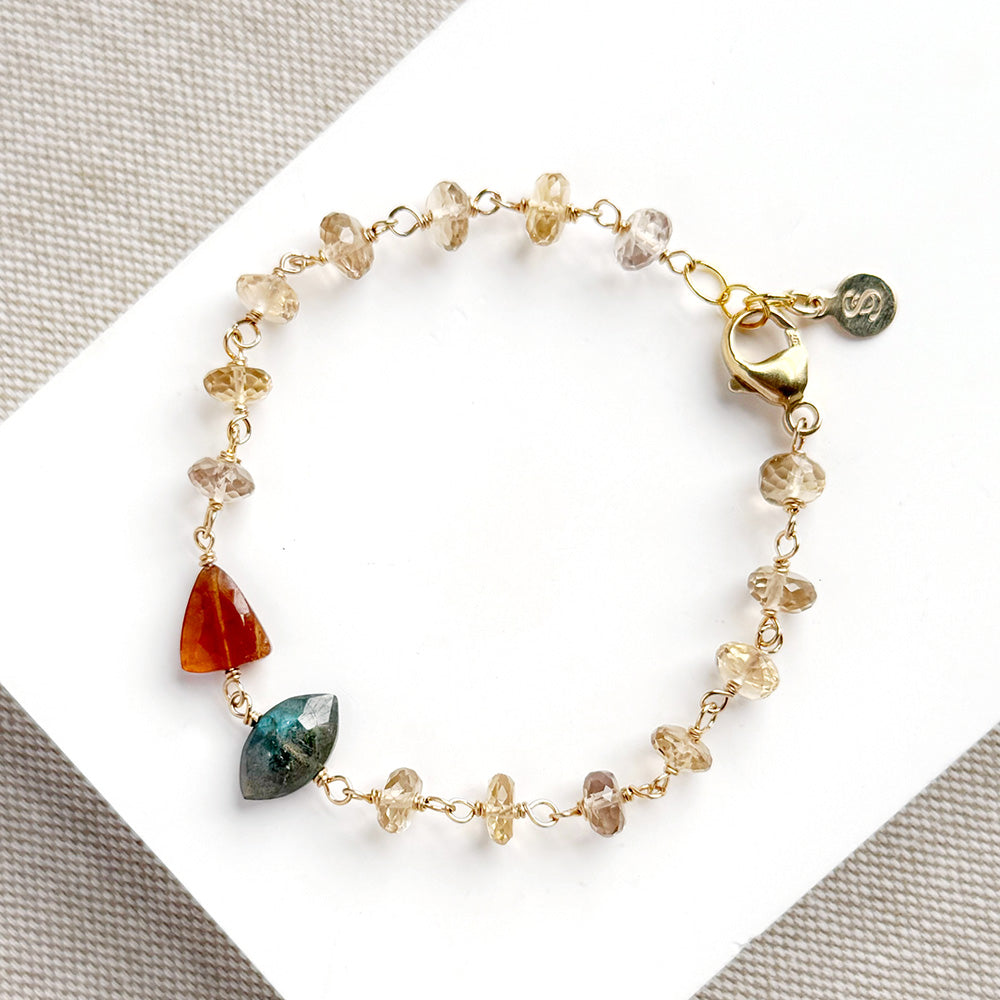 A gold chain bracelet with small faceted clear and beige beads, a triangular amber-orange stone, and a marquise-cut green stone, displayed on a white card over a textured gray fabric background.