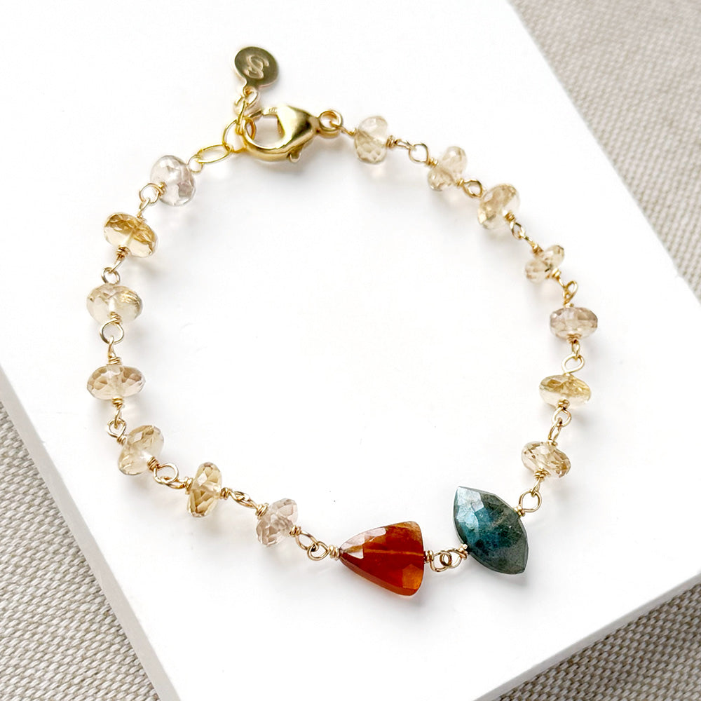 A delicate gold chain bracelet with clear faceted crystal beads and two prominent gemstone charms, one orange-red and one teal-green, displayed on a white card.