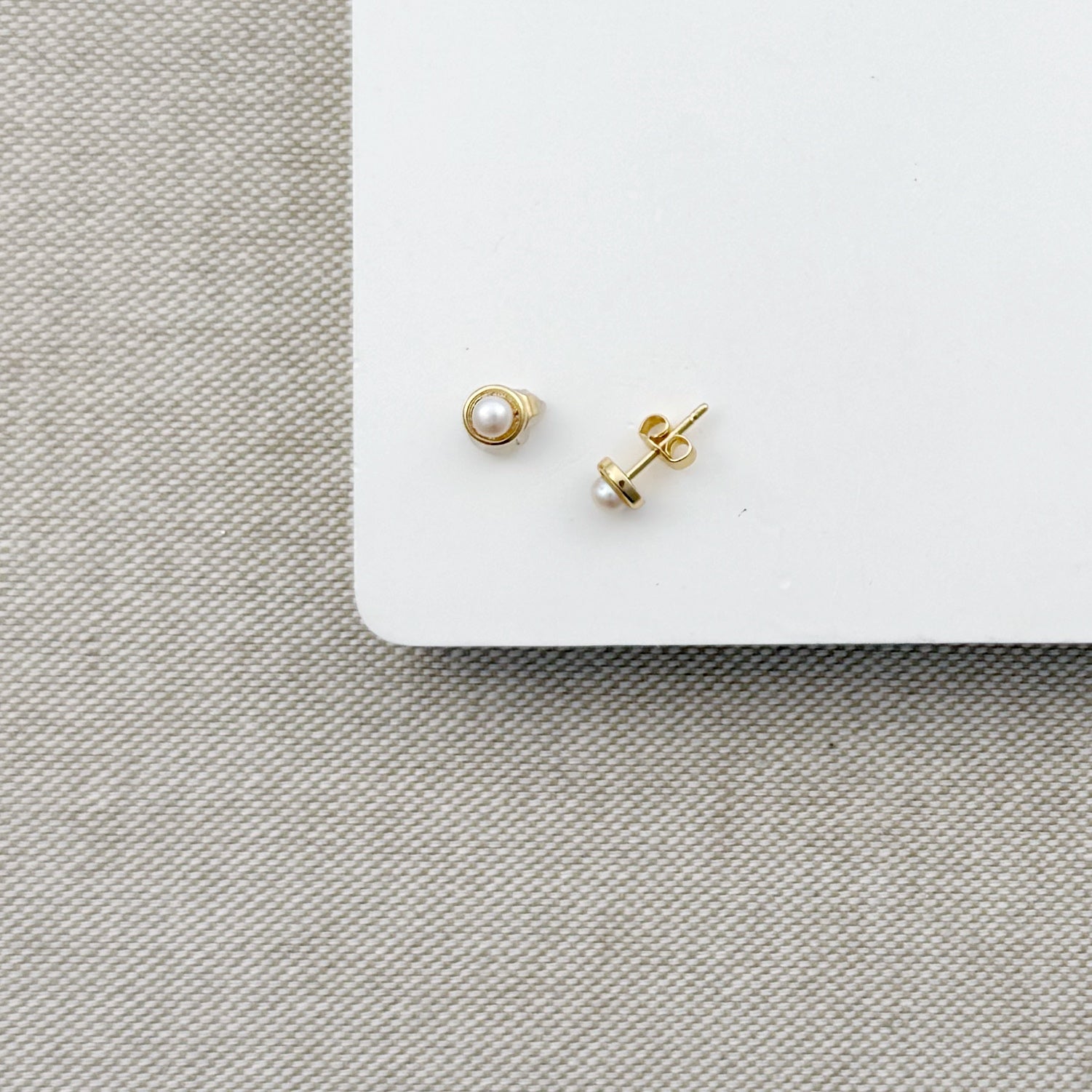 Pair of gold and pearl earrings on a white square platform with a gray fabric background