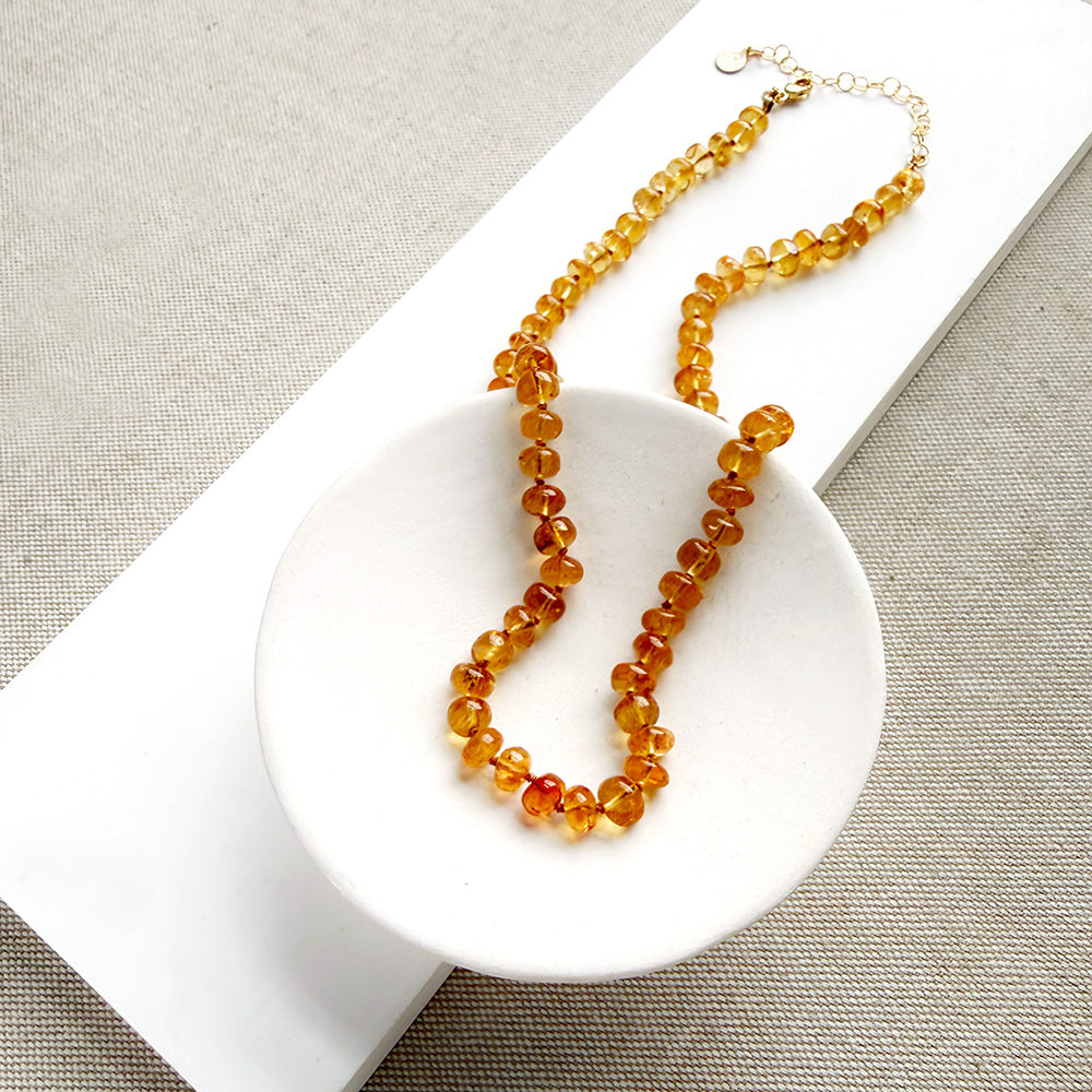 A beaded necklace made of smooth Imperial Topaz with a gold clasp, draped over a small white dish on a textured beige surface.