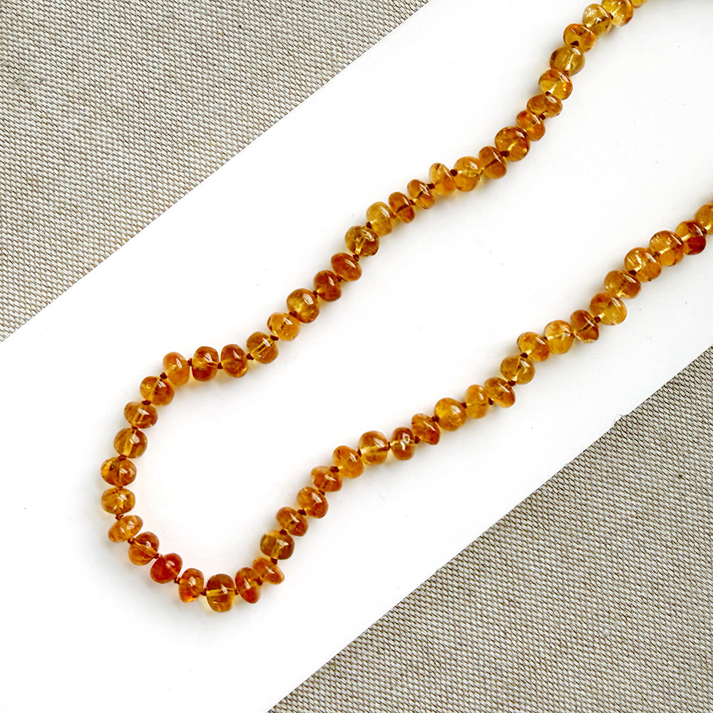 A beaded necklace made of smooth Imperial Topaz with a gold clasp, draped over a small white dish on a textured beige surface.