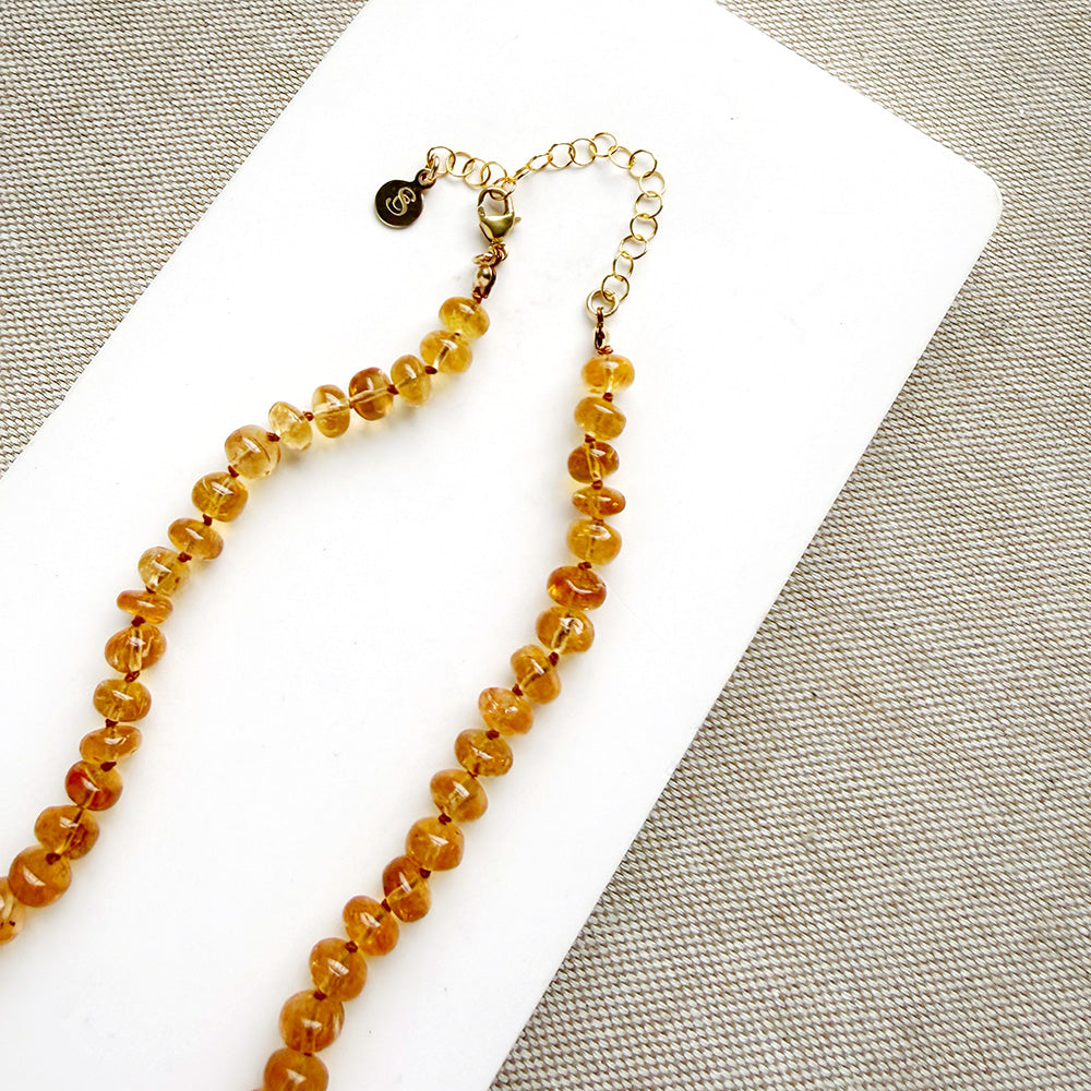 A beaded necklace made of smooth Imperial Topaz with a gold clasp, draped over a small white dish on a textured beige surface.