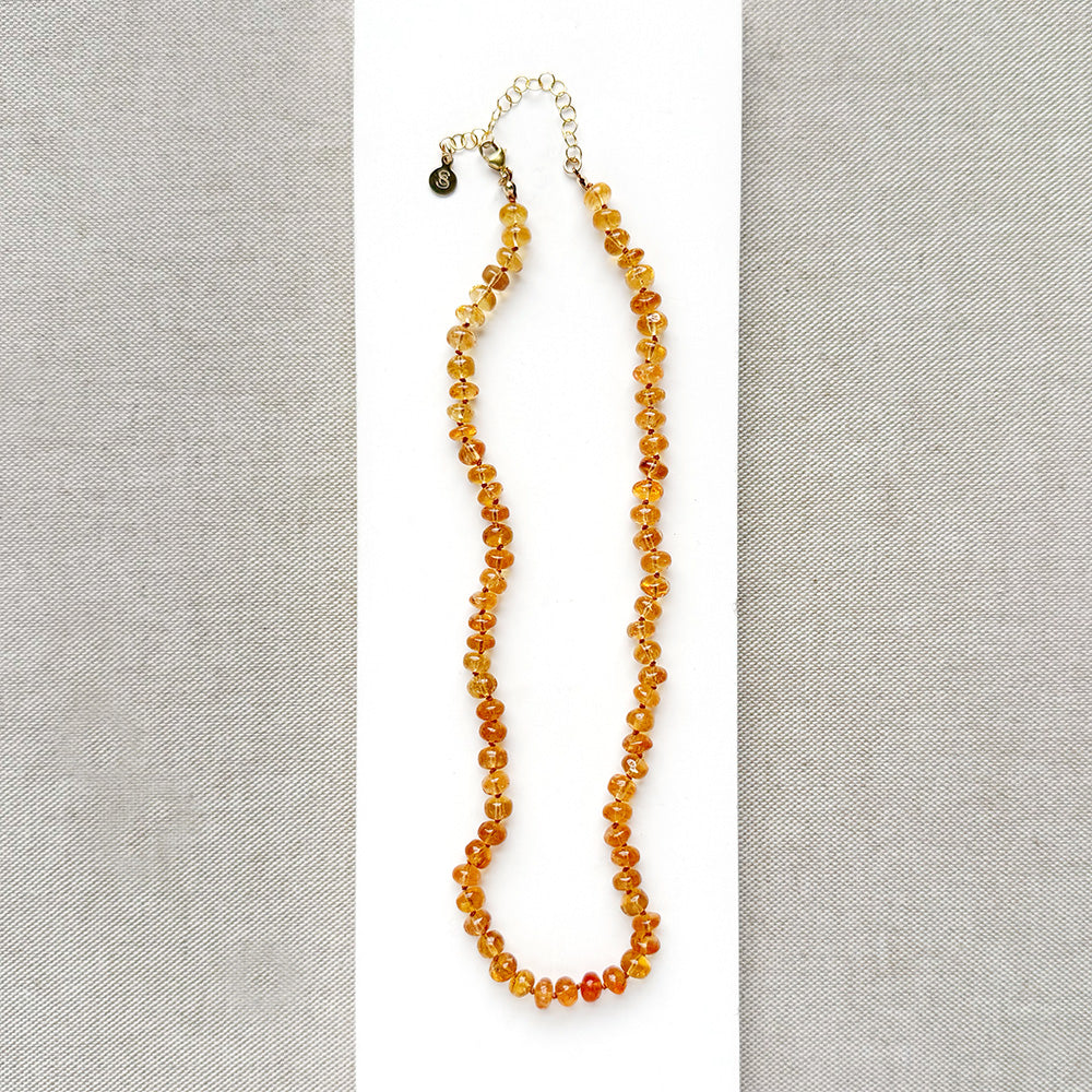 A beaded necklace made of smooth Imperial Topaz with a gold clasp, draped over a small white dish on a textured beige surface.