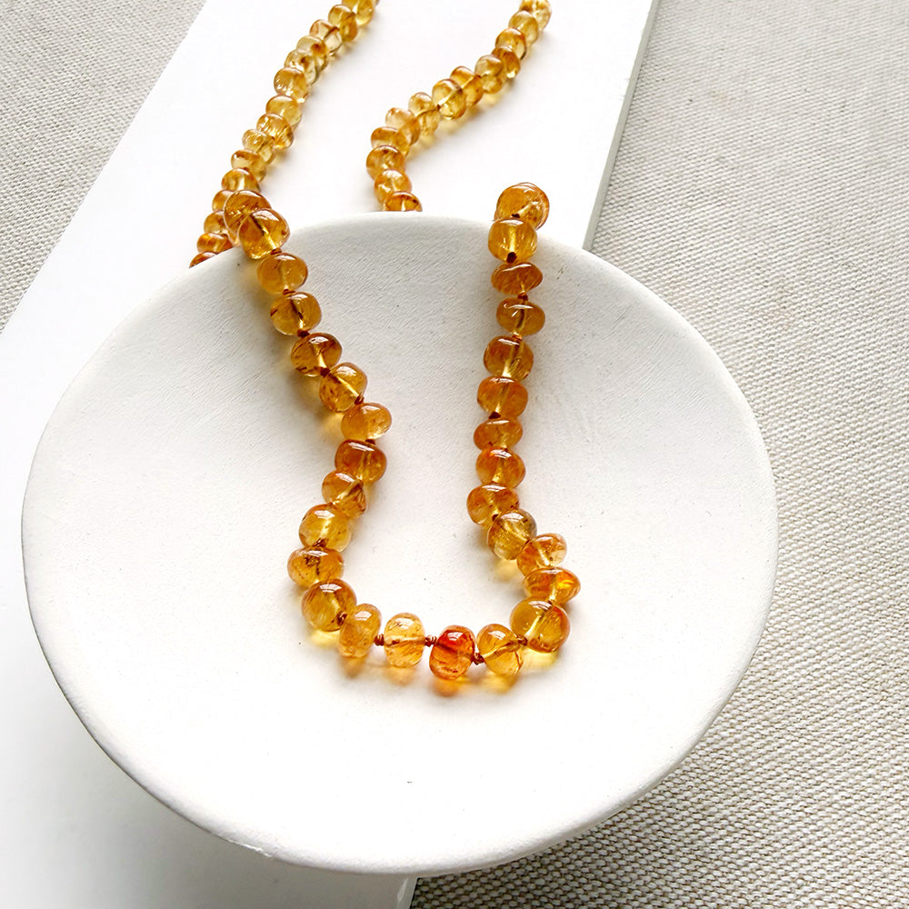 A beaded necklace made of smooth Imperial Topaz with a gold clasp, draped over a small white dish on a textured beige surface.
