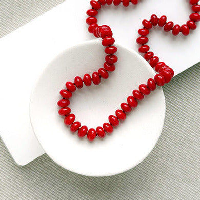 A coral knotted necklace in a rich red color with a gold clasp, displayed on a white ceramic dish over a light gray textured fabric background.