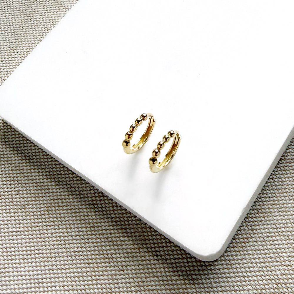 A pair of small gold hoop earrings with a textured beaded edge, displayed on a white square card over a woven beige surface.