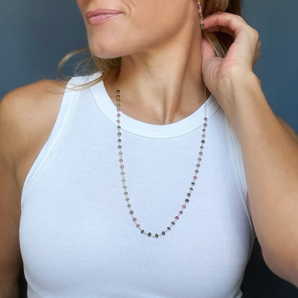 A woman wearing a delicate beaded necklace with watermelon tourmaline gems, paired with a white ribbed tank top against a dark blue-gray background.