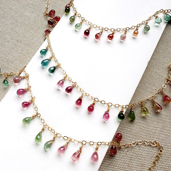 Gold necklaces with dangling teardrop-shaped Watermelon Tourmaline gemstones in pink, green, and teal, displayed on a white card over a beige textured fabric background.