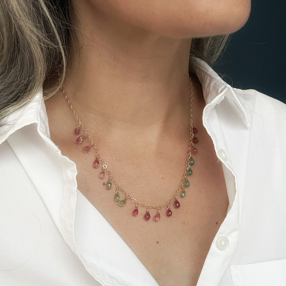 A close-up of a woman wearing a delicate gold necklace with small dangling Watermelon Tourmaline teardrop gemstones in pink, teal, and green, styled with a white collared shirt.