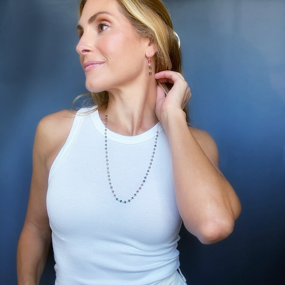 A woman wearing a delicate gold beaded necklace and matching drop earrings, posing against a dark blue gradient background.