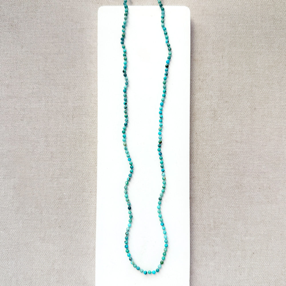 A long beaded necklace made of small turquoise-blue beads with a few dark accents, displayed on a white rectangular card against a textured light gray fabric background.