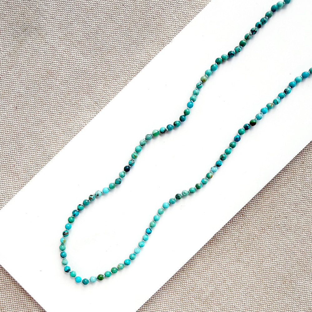 A beaded strand necklace made of small turquoise-blue and green marbled beads, laid diagonally on a white rectangular card over a beige woven fabric background.