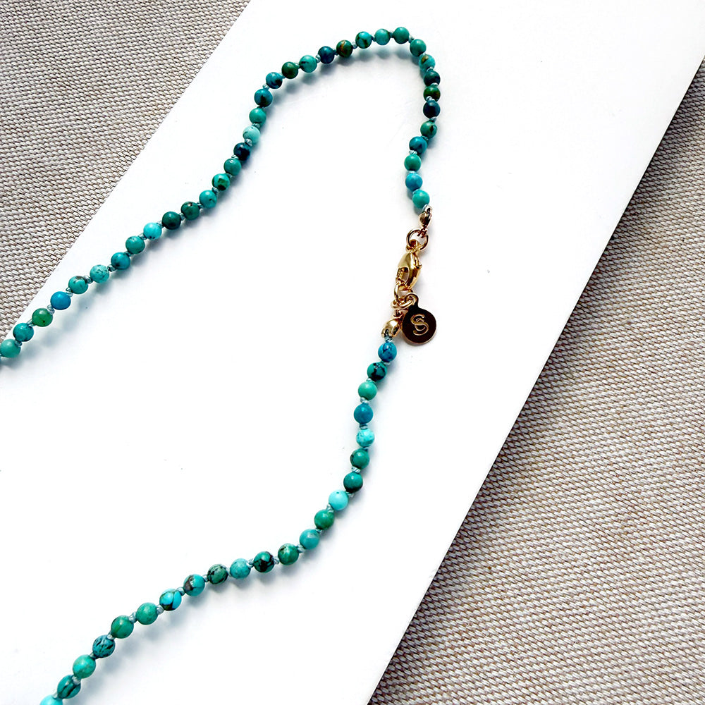 A beaded necklace made of small turquoise and green beads with a gold clasp and a small round charm, laid on a white card over a textured gray fabric background.