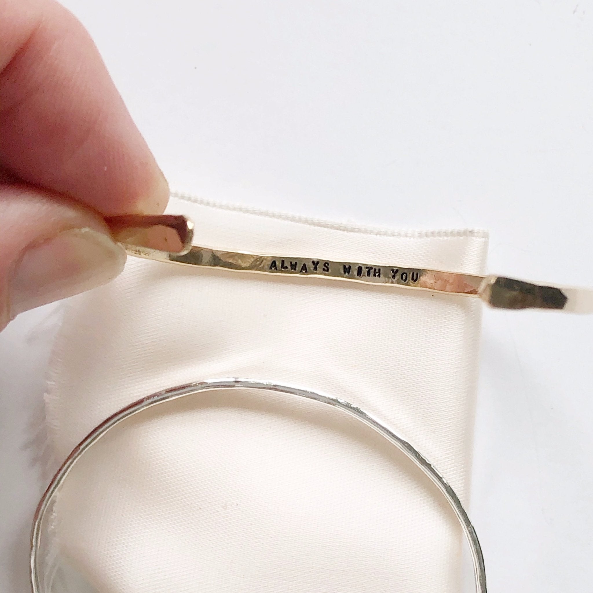 2 gold and silver textured bangle bracelets with the words "always with you" stamped on the inside. Fingers holding one bracelet. Always with You Bangle by Sarah Cornwell Jewelry