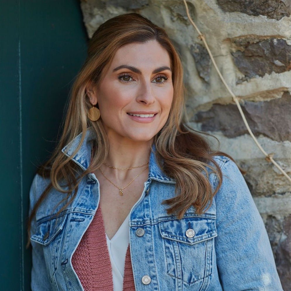 Gold Buffay Earrings by Sarah Cornwell Jewelry. Blonde woman wearing denim jacket, pink sweater, lightweight, shimmery, textured 1 inch gold disc earrings stacked with gold studs and gold layered necklaces on stone background.