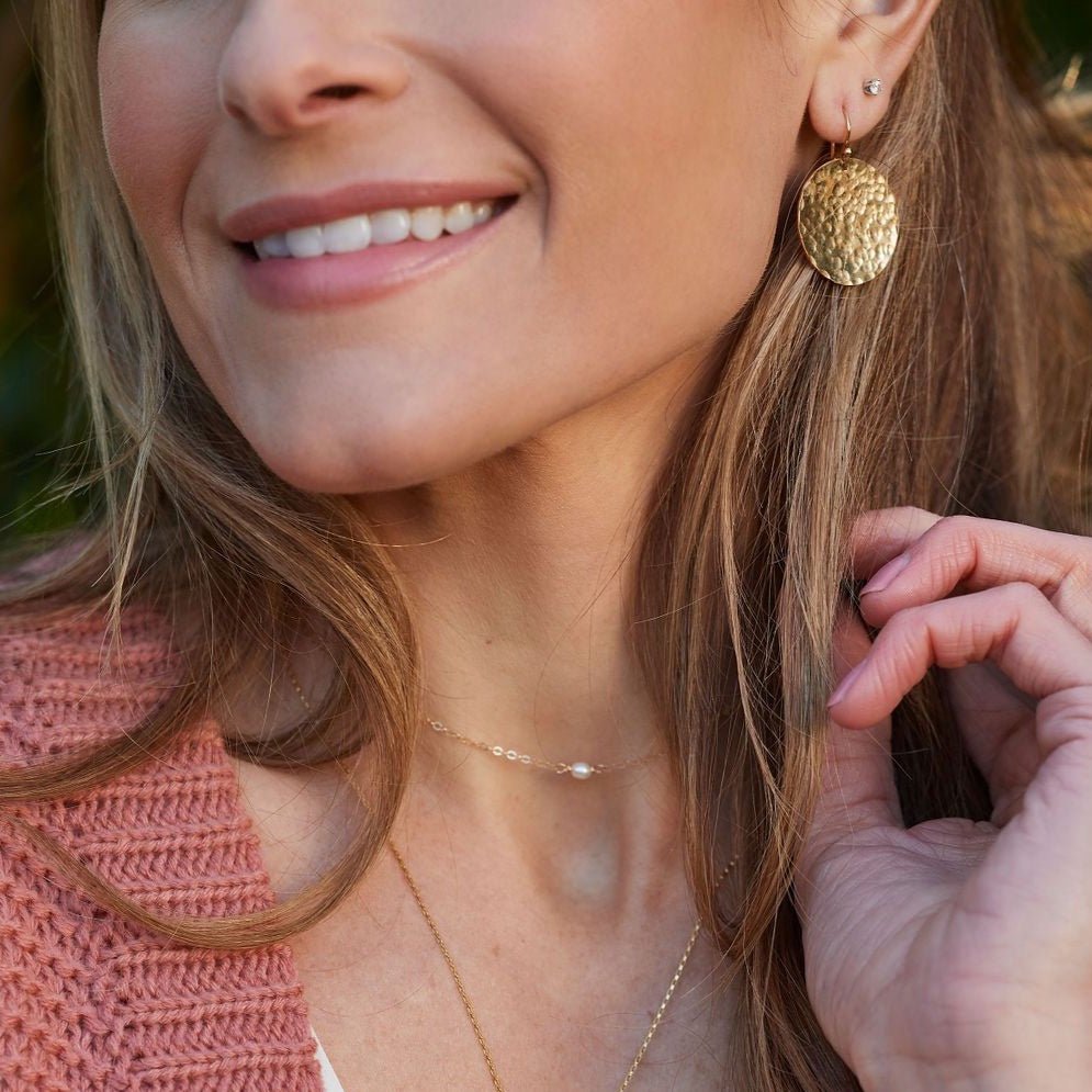 Gold Buffay Earrings by Sarah Cornwell Jewelry. Blonde woman wearing pink sweater, lightweight, shimmery, textured 1 inch gold disc earrings stacked with gold studs and gold layered necklaces.