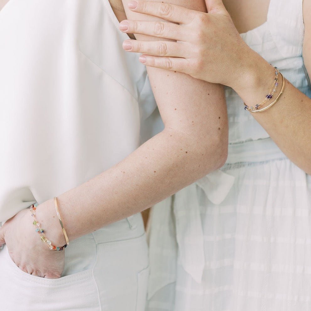 Close up of 2 women's arms wearing all white with gold textured bangle bracelets and gold and multicolored gemstone bracelets. Caroline Bangle by Sarah Cornwell Jewelry