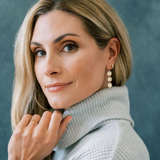 Side view of a woman with blonde hair wearing a gray sweater with gold 1.5 inch drop earrings with 3 same sized textured discs and gold huggie hoops in her second piercing. Cole Earrings by Sarah Cornwell Jewelry