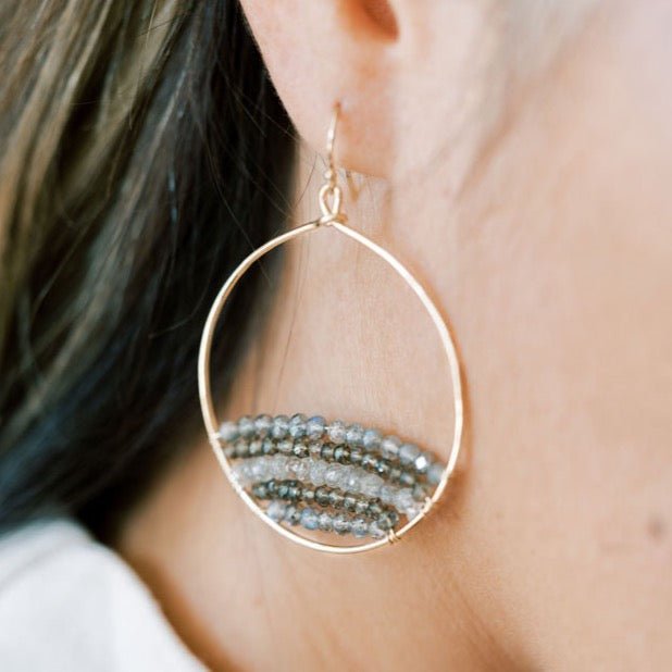Close up of woman with long dark hair wearing a white shirt with gold and gemstone statement 2 inch hoop earrings with rows of zircon, labradorite, and smokey topaz gemstones. Harper Earrings by Sarah Cornwell Jewelry